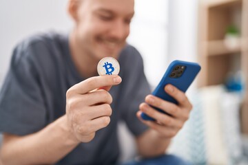 Young caucasian man using smartphone holding bitcoin at home