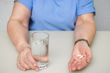 Senior woman sitting at the table and holding in the old wrinkled hands white capsules, vitamins or pills for treatment and water glass, healthcare and medicine concept