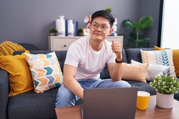 Young asian man using laptop at home sitting on the sofa smiling looking to the side and staring away thinking.