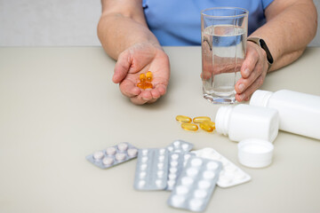 Senior woman with wrinkled old hands at the table holding omega 3 yellow capsules, fish oil pills and water glass. Healthcare and medicine concept