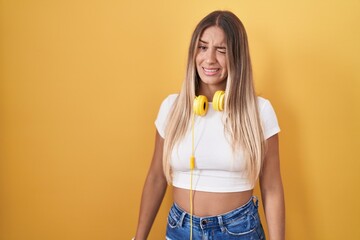 Young blonde woman standing over yellow background wearing headphones winking looking at the camera...