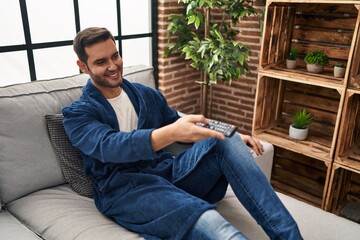 Young hispanic man watching tv sitting on sofa at home