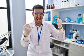 Young hispanic man with beard working at scientist laboratory holding blue ribbon annoyed and frustrated shouting with anger, yelling crazy with anger and hand raised