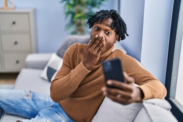Young african man with dreadlocks using smartphone doing video call covering mouth with hand,...