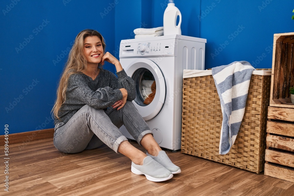 Wall mural young blonde woman sitting on the floor waiting for washing machine at laundry room