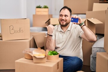 Plus size hispanic man with beard moving to a new home holding credit card pointing thumb up to the side smiling happy with open mouth