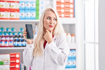 Caucasian woman working at pharmacy drugstore showing smartphone screen serious face thinking about question with hand on chin, thoughtful about confusing idea
