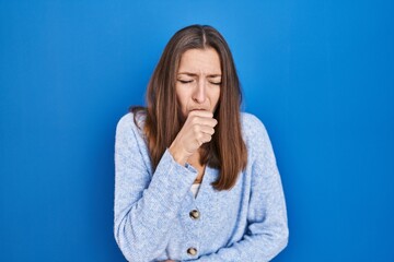 Young woman standing over blue background feeling unwell and coughing as symptom for cold or...