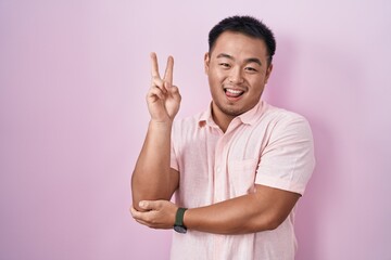 Chinese young man standing over pink background smiling with happy face winking at the camera doing victory sign with fingers. number two.