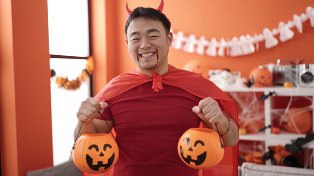 Young chinese man wearing devil costume holding halloween pumpkin baskets at home
