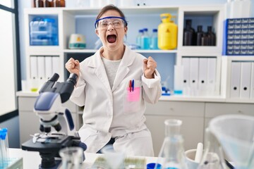 Hispanic girl with down syndrome working at scientist laboratory celebrating mad and crazy for success with arms raised and closed eyes screaming excited. winner concept