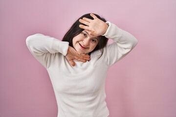 Woman with down syndrome standing over pink background smiling cheerful playing peek a boo with hands showing face. surprised and exited
