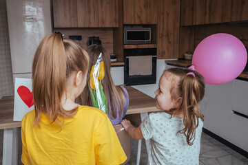Daughters congratulate their mom on Mother's Day, a card with a heart, flowers and a balloon at home in the kitchen. Children surprise their mother for the holiday.