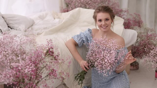A beautiful woman in a dress sits on the floor in the bedroom next to the bed among bouquets of pink gypsophila flowers. She is holding a bouquet in her hands.
