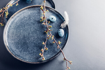 Easter dish decorated with blue eggs, feathers and spring branches. Ceramic plate on grey background. Space