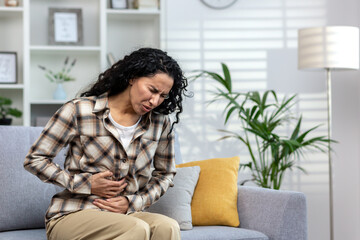 Sick mature adult woman sitting on sofa alone at home, having severe stomach pain, Latin American...