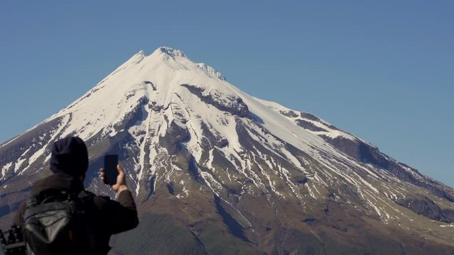 Majestic Mount Taranaki and young man with face mask taking picture with smartphone. Selective focus