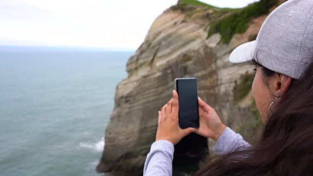 Woman filimig with mobile at amazing cliff landscape at Cape Farewell, New Zealand