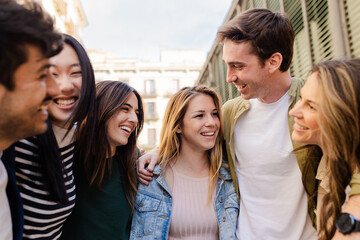 Friendly group of young friends laughing together in city street. United millennial people hugging each other while hang out having fun on vacation