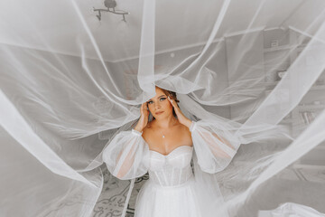 Beautiful curly brown-haired bride in a white dress poses for a photographer, standing under a veil in a beautiful dress with sleeves. Wedding photography, close-up portrait, chic hairstyle.