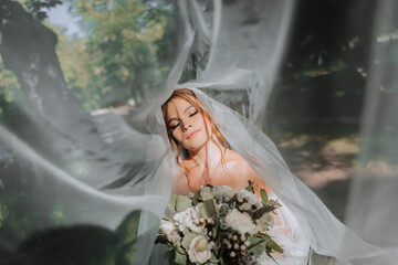 Beautiful curly brown-haired bride in a white dress poses for a photographer, standing under a veil...