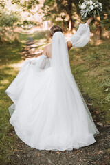 A beautiful bride in a white dress with a long veil walks away from the camera with a bouquet in the park. Photo from the back. Young woman, art photo, wedding, bride, open shoulders. light.