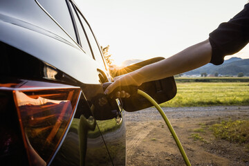 Female hand plugging in a charger in opened charging socket on a black electric car, with the sun...
