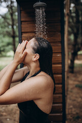 A girl takes a shower with fresh water on the beach or the street after sunburn, surfing and the sea Beautiful body, summer mood. Brunette in black swimwear enjoying natural water 