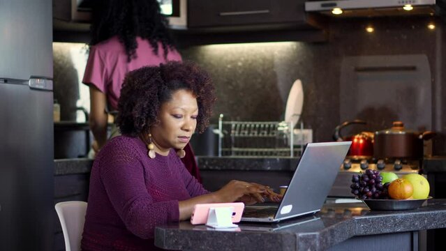 Black woman working with laptop and mobile in the kitchen. Mother typing and daughter washing dishes.
