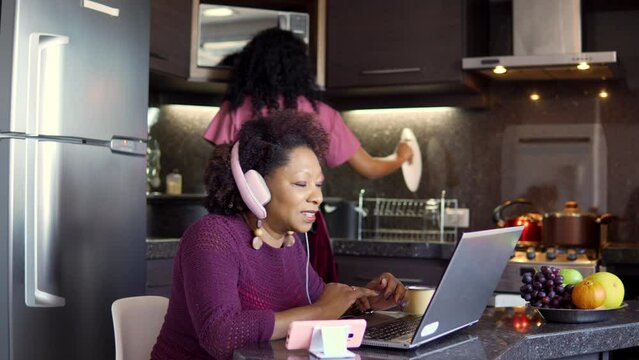 Black woman in video call with headphones and laptop. Girl washing dishes and cooking in the back.