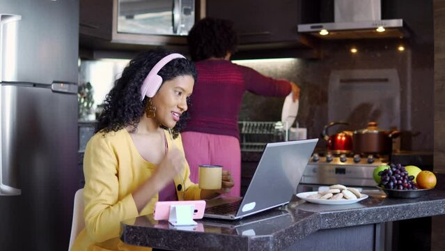 Black woman in video call and mother busy cooking in the kitchen. Young student talking online with headphones.