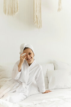 cheerful girl sitting on the bed in a bathrobe applies cream on her face. sexy girl after shower. body care. dermatology. face treatment