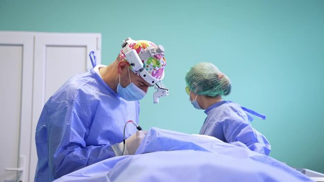 Mid-aged male surgeon with headlight uses electric device instrument. Female nurse assisting the professional.