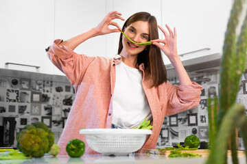Happy Young beautiful cheerful girl making mustache from green bean on face at home kitchen and smiles. The concept of a healthy nutrition and vegan lifestyle. Green vegetables. Real emotions
