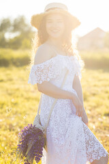 Fototapeta na wymiar Beautiful young girl in a white dress, straw hat, with picnic and bouquet of purple wild flowers on a meadow. Summertime, golden hour, sunset. Wellness, wellbeing. Calendar