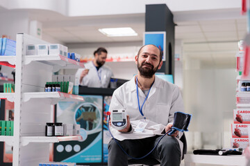Portrait of pharmacist holding medical tonometer ready to measure clients hypertension during checkup visit in pharmacy. Drugstore filled with supplements and vitamins, medicine service