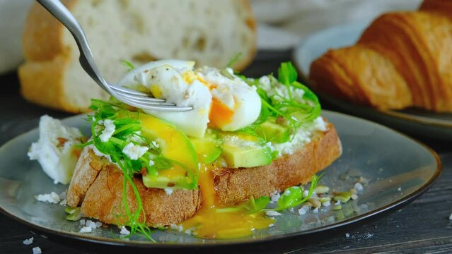 Person Eating A Poached Egg On Avocado Toast, Cutting A Benedict Egg With Avocado Spread On Sandwich With Fork, Runny Egg Yolk, Healthy Breakfast, Close-up Video Clip, 4k High Quality Footage