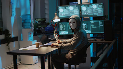 Male criminal wearing mask and hood to hack computer system, breaking into company servers to steal big data. Masked man looking dangerous and scary, impostor creating security malware. Handheld shot.