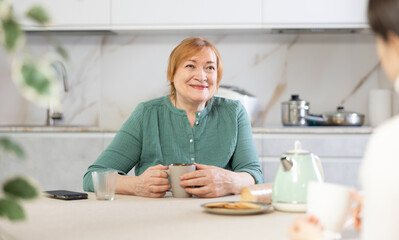 Cheerful adult woman and young woman in casual clothes drinking tea in kitchen