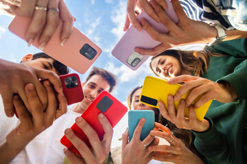 Young group of people using mobile phone device standing together in circle outdoors. Millennial friends addicted to social media app, betting or playing video game on platform online. - obrazy, fototapety, plakaty