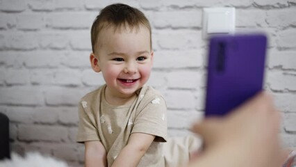 Beautiful smiling Caucasian kid looks at phone. Parent showing video on the smartphone to a baby boy. White wall backdrop.