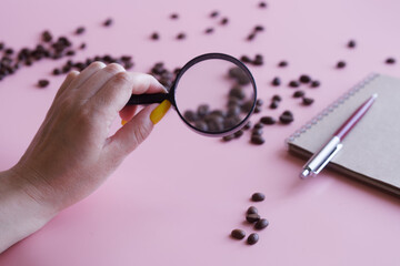 Woman's hand holds magnifier next to coffee beans, notepad and pen. Quality of roasted coffee beans. Examination coffee quality. Analysis of the content of useful substances. Selective focus.
