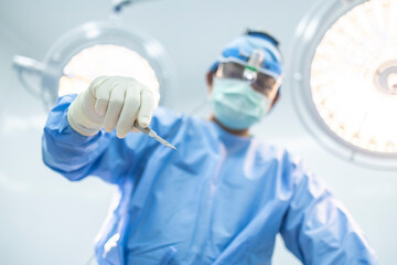 Uprisen of surgeon or doctor holding scalpel inside operating room in hospital with blurred surgical lamp background.Surgeon in blue uniform did surgery technology.Selective focus at hand of people. - obrazy, fototapety, plakaty