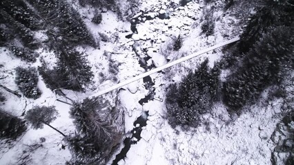 Fast dramatic aerial shot of hanging bridge ovr a small river in the middle of a snow covered forest.