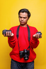Young Asian man in casual style with dslr camera confuse to choice lens against yellow background. 