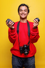 Handsome young Asian man in casual style with dslr camera smiling while holding lenses against yellow background. 