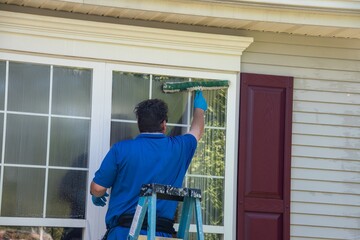 A Caucasian man on a ladder wearing blue latex gloves and listening to ear buds washing a window with a squeegee © ALAN