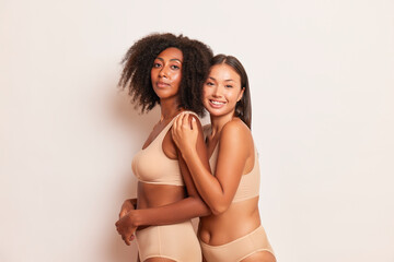 Two lovely model girls in beige underwear are standing embracing on white background, happy African American and Asian young woman, friendship concept, copy space, high quality photo