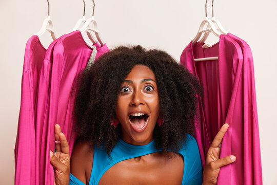Cheerful African American young woman is standig between pink dresses hanging on clothes rail, happy about her shopping, shopping time concept, copy space, high quality photo