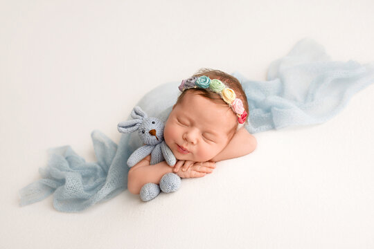 A newborn baby girl in a flower headband sleeps with a blue rabbit in her hands. The girl lies on white background covered with a light blue blanket.